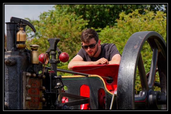 A man wearing sunglasses and a t-shirt leans over a large, vintage steam engine. The engine is black and red with gold accents, and a large black flywheel is visible on the right. In the background, green trees are out of focus under a bright sky. The man appears to be inspecting or resting on the machinery.