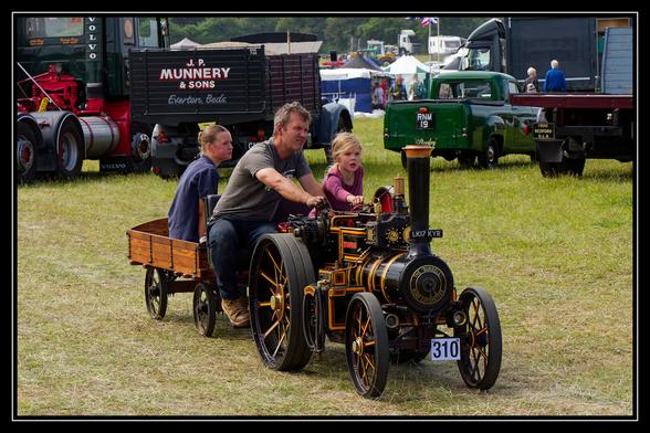 A man and two young girls are riding on a miniature traction engine in a grassy field. The man is seated in front, operating the engine, with one girl directly behind him and the other in a small wooden trailer attached to the back. The traction engine is black and gold with intricate details. In the background, other vintage vehicles, including trucks and a green pickup, are parked on the field under a cloudy sky, suggesting an outdoor show or event.