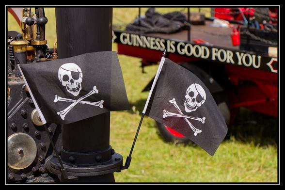 Two small pirate flags, each featuring a white skull and crossbones on a black background, are displayed in a grassy outdoor setting. One flag is attached to a dark metal pole, possibly part of machinery, in the foreground. The other flag is slightly further back and to the right. In the background, a red and black vehicle or trailer is visible with the words "GUINNESS IS GOOD FOR YOU" painted on its side, partially obscured. The ground is covered in green grass, and the overall scene suggests an outdoor event or display.