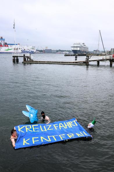 Drei Menschen schwimmen in der Kieler Förde. Sie halten ein Schwimmtranspi auf dem Kreuzfahrt kentern steht. Ein Mensch hält einen aufblasbaren Plastikwal. Im Hintergrund ist ein Kreuzfahrtschiff und die Stena Line zu sehen