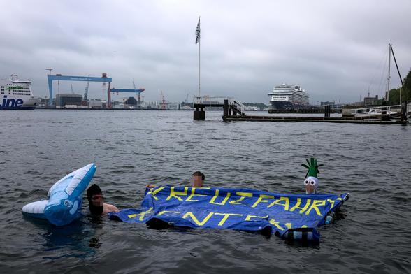 Drei Menschen schwimmen in der Kieler Förde. Sie halten ein Schwimmtranspi auf dem Kreuzfahrt kentern steht. Ein Mensch hält einen aufblasbaren Plastikwal. Im Hintergrund ist ein Kreuzfahrtschiff und die Stena Line zu sehen