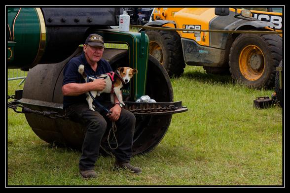 An older man wearing a black polo shirt and a "JCB" baseball cap is sitting on the front roller of a vintage green and black steamroller, holding a small terrier dog. The dog is white and brown with a pink harness. In the background, a large yellow JCB construction vehicle is partially visible on the right, and the setting appears to be a grassy field, possibly at an outdoor show or event.