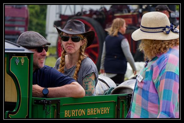 Three people are gathered around a green miniature train carriage with "BURTON BREWERY" written on its side. On the left, a man in a flat cap and sunglasses is smiling. In the center, a woman with braided hair, sunglasses, and a cowboy hat looks directly at the camera with a slight grimace, revealing colorful tattoos on her arm. On the right, a woman in a straw hat with a blue ribbon and a colorful plaid shirt is seen from behind, facing the other two. In the background, other people and parts of large machinery are visible, suggesting an outdoor event.