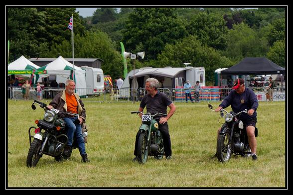 Three men are seated on vintage motorcycles in a grassy field, seemingly at an outdoor event. The man on the left, with an orange jacket and beard, is on a white motorcycle. The man in the middle, wearing a dark shirt, is on a green motorcycle with the number "282" visible. The man on the right, wearing a red cap and dark jacket, is on a black motorcycle. In the background, there are tents, caravans, and trees under a cloudy sky, with people milling about. A Union Jack flag is visible on a flagpole in the far background.