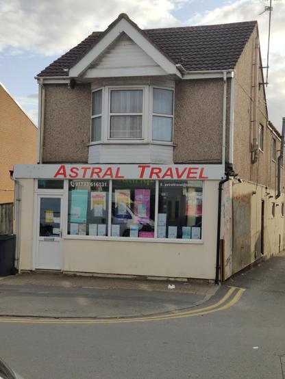 A grim looking 1980s building which is a shop front topped with a plastic window framed bay window on the second story and a peaked dormer roof on top of that, with white plastic shuttering. The sign over the shop front says "ASTRAL TRAVEL" in generic red plastic sans serif italic capital letters.