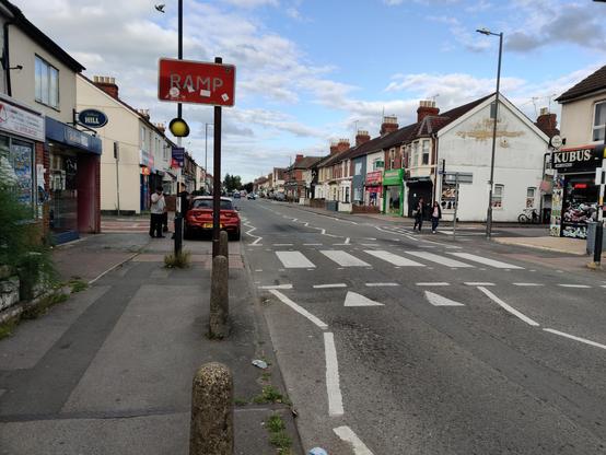 A view up a nondescript stroad in Swindon looking north. This street is just north of the train station and to the east of the historic works site. A zebra crossing is visible in the foreground. The buildings are two-story Victorian terraces, with shops interspersed among bay windowed residential houses with pitched tiled roofs and three pot brick chimneys.