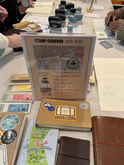 The image shows a table set up for a stamp-related event, with a sign in the center that reads "STAMP CARAVAN USA 2025." The sign features a map and lists various locations and dates, including "The Paper Mouse (Norton, MA)" from May 1-5, 2025, and "Penny Post (Alexandria, VA)" from May 29-31, 2025. The table is covered with various stationery items, including notebooks, stamps, and stamp pads. There are several notebooks with different covers, some featuring stickers and designs, such as a brown notebook with a suitcase sticker and a map of the San Francisco Bay Area. The notebooks are arranged in a semi-circle around the sign. In the background, people are seen handling stamp pads and other stationery items, suggesting an interactive event. The overall setting appears to be a craft or stationery fair, with a focus on stamp collecting and related activities.

Provided by @altbot@fuzzies.wtf, generated privately and locally using Ovis2-8B

🌱 Energy used: 0.235 Wh
