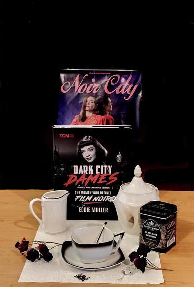 Tea tableau in a chiaroscuro mood featuring artwork from Eddie Muller's Noir City Festival and his book Dark City Dames. The tea service elements are MidCentury pieces from the 1940s, and to pound home the theme there is a black spider made of faceted black and purple jewels, a tiny silver handmirror shaped like a heart and edged in sparkly diamonds, and bouquets of red roses, except the blossoms are dried out, appearing the color of darkening blood. The tea is Black Cask Bourbon black loose-leaf from Harney and Sons.