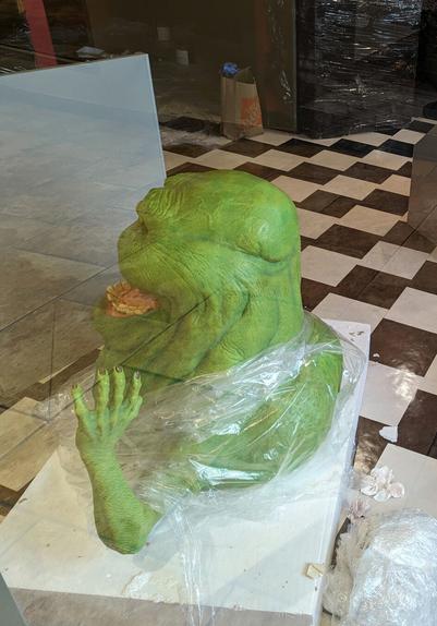 A "life"-sized Slimer from Ghostbusters emerging from plastic wrap on a white platform behind glass. Floor is checkered
