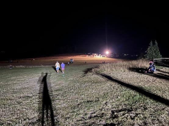 Campingwiese in der Nacht. Langer Schlagschatten einer Person unter einem Flutlicht. Zwei Personen kommen entgegen. Weit im Hintergrund ein Lichtkegel eines anderen Flutlichts. Dazwischen und daneben: Dunkelheit.