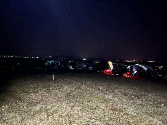 Braun grüne Wiese von einem Lichtkegel erhellt. In der Mitte Campingfahrzeuge und Zelt mit diffusen bunten Lichtquellen. Im Hintergrund beleuchtet Ortschaften