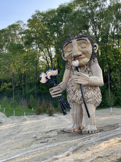 Troll made of wood sniffs street lamp as though it were a flower while holding a bouquet of street lamps in the other hand.