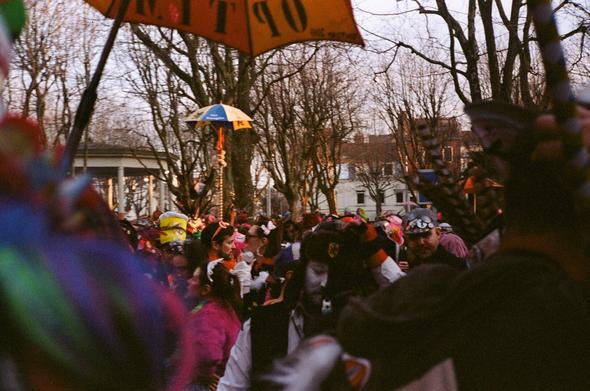 Colored photography of a crowd of people disguised. in the middle of the photography (and in the foreground) there are umbrella with a very long handle). there are leaveless trees and buildings in the background.
