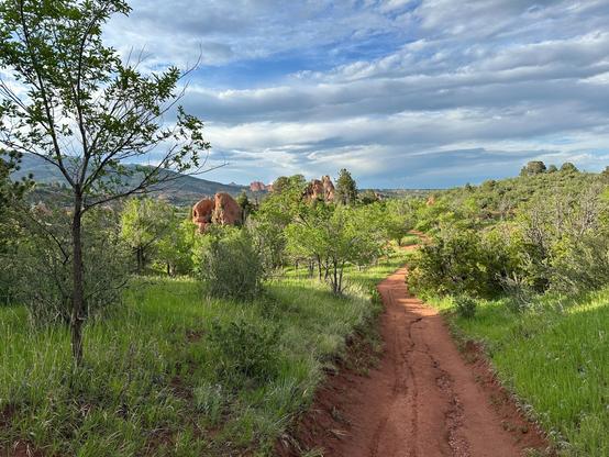 A trail wends up throug the center-right side of the image. Red sandstone rock formations pop up through the greenery on the left. Partly cloudy skies overhead.