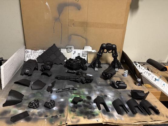 A makeshift airbrush area, made of cardboard on a wooden work bench. A collection of reaver Titan pieces litters the cardboard. All are black. A piece of styrofoam is to the right, with little pieces on legs. Also black. A piece of terrain is leaning against the cardboard. Also black.