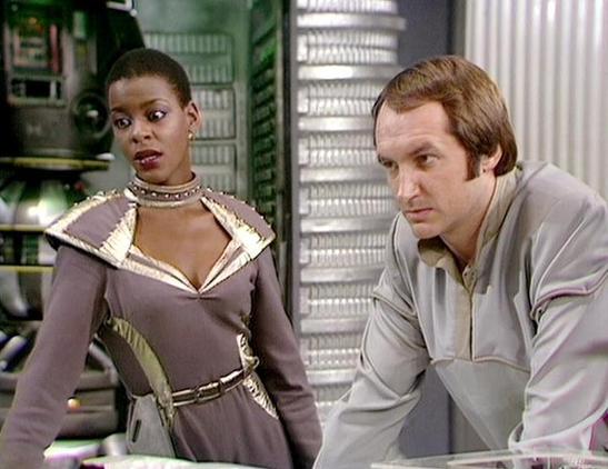 Claude Sonnet 4.0 describes the image as: "This appears to be a scene from a science fiction television series, showing two characters in what looks like a spacecraft or futuristic facility interior. The setting features typical sci-fi production design with metallic walls and technical equipment visible in the background. One character is wearing an elaborate costume with metallic trim and ornate detailing around the collar and belt, while the other is dressed in more utilitarian clothing. The lighting and overall aesthetic are characteristic of British science fiction television from the late 1970s or early 1980s era."

Blakes7Bot, version 3.9.30.