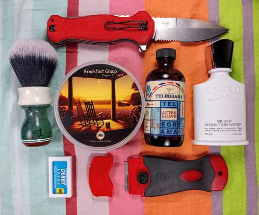 (Le Chat Mistral description)

The image displays a collection of items arranged on a striped background. At the top, there's a red folding knife with a red handle. To the left, there's a shaving brush with white and black bristles. Below the brush, there's a small blue and white pack of razor blades labelled "Derby Extra."

In the centre, there's a round tin with an image of a porch and rocking chairs, labelled "Breakfast Group." To the right of the tin, there's a bottle of aftershave with a vintage label reading "Telegrama After Shave." Next to it, there's a white bottle of "Creed Silver Mountain Water" cologne.

At the bottom, there's a red plastic item, possibly a razor handle, and a red and black razor with a blade attached.