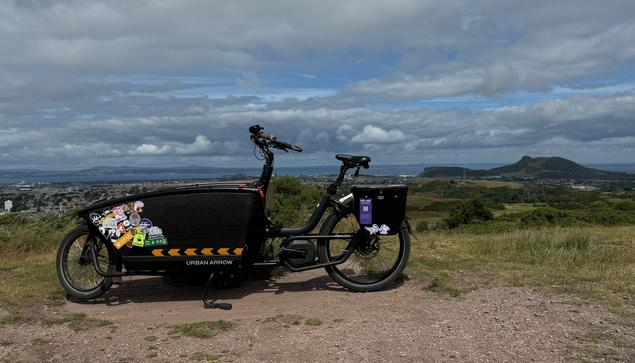 The weather is partly sunny; under a mix of cloud and blue skies, a black, bestickered, front-loading cargo bike is parked at the top of a hill with a sprawling view of Edinburgh behind it. To the right of frame, the prominent figure of the crags and Arthur’s Seat loom up in the middle of the otherwise urbanised view; in the background, the sea and hills beyond.