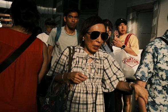 A scene from a crowded footpath. An individual wearing sunglasses clasps the arm of another individual. Another person directly behind them to the left looks above and past the camera. A third person behind the individual in sunglasses holds a phone to their ear looking the right of the photo while wearing a ball cap and holding a bag that says "Marque Déposée Danton France". Other people are partially visible in the frame walking towards and away from the camera.