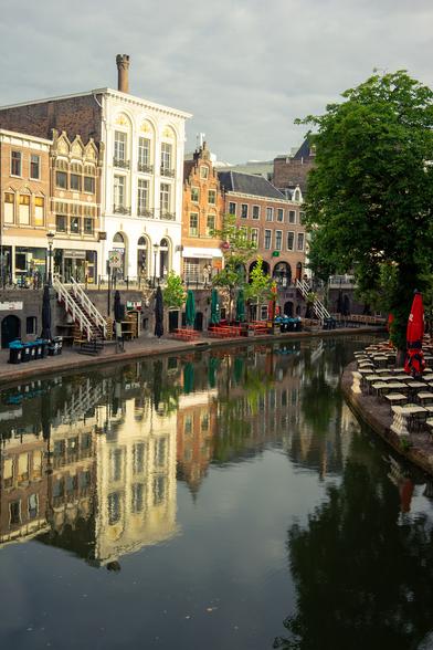 The image depicts a serene canal scene in a European city, likely in the Netherlands, characterized by its historic architecture and waterways. The foreground features a calm canal with reflections of the surrounding buildings, creating a mirror-like effect. On the left side, a row of traditional buildings with brick facades and ornate details, including arched windows and decorative elements, line the canal. The buildings have a mix of white and brown tones, with some displaying commercial signage. The right side of the canal shows a tree-lined promenade with outdoor seating, including tables and red umbrellas, suggesting a café or restaurant area. The sky is overcast, adding a soft, diffused light to the scene. The overall atmosphere is peaceful and picturesque, with the still water enhancing the symmetry of the composition.

Provided by @altbot, generated privately and locally using Ovis2-8B

🌱 Energy used: 0.198 Wh