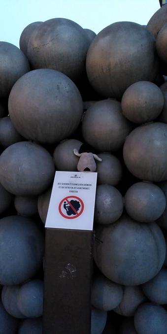 Archy klettert auf einem Kunstwerk, das eine Mauer aus verschieden großen Steinkugeln ist. Unter Archy ist ein Schild sichtbar mit der Aufschrift "Aus Sicherheitsgründen ist das Klettern am Kunstwerk verboten"