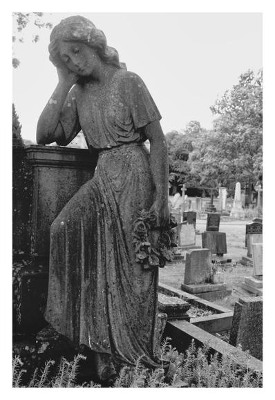 A weathered stone statue of a woman stands in a quiet cemetery, leaning gently on a pedestal with one arm propping up her bowed head. Her expression is serene and sorrowful, eyes closed as if deep in thought or mourning. She wears a long, flowing gown, and in her lowered hand she holds a bouquet of carved flowers, worn by time. Gravestones stretch into the background, softened by trees and overcast sky. The image is in black and white, enhancing the solemn, timeless atmosphere. Moss and age spots mark the statue, suggesting long years spent watching over the resting souls around her.