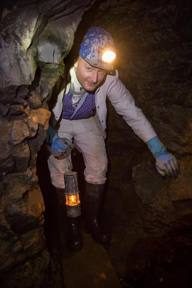 Farbfoto eines Bergmanns in einem engen Stollen, der eine Grubenlampe trägt und einen Schutzhelm mit Stirnlampe auf dem Kopf hat. Er trägt Arbeitskleidung, Gummistiefel und blaue Handschuhe, während er sich vorsichtig durch den schmalen Tunnel bewegt.