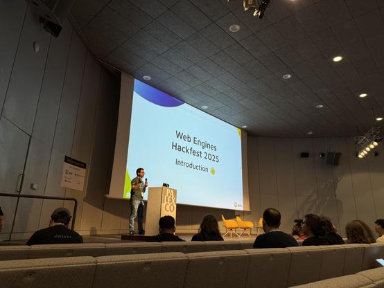A man standing in front of podium on a stage in a large auditorium. Behind him, a screen projects a slide that reads "Web Engines Hackfest 2025 - Introduction"