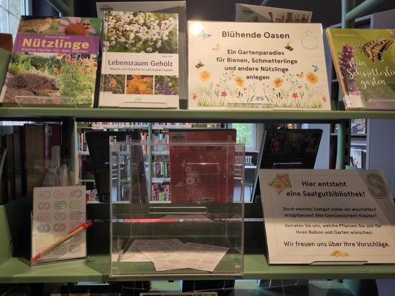 Das Bild zeigt ein Regal in der Bezirkszentralbibliothek Spandau mit einem Display zum Thema Gärtnern und Natur. In der oberen Reihe sind mehrere Bücher ausgestellt, darunter Titel wie "Nützlinge: Kleine Helfer für den Garten", "Lebensraum Gehölz: Bäume und Sträucher im naturnahen Garten". Oben ist ein Schild mit dem Text: "Blühende Oasen: Ein Gartenparadies für Bienen, Schmetterlinge und andere Nützlinge anlegen".

In der unteren Reihe, auf der Regalfläche, steht prominent eine durchsichtige Sammelbox. Daneben befindet sich ein Schild mit der Aufschrift "Hier entsteht eine Saatgutbibliothek!". Darunter steht ein Text auf Deutsch, der die Besucher auffordert, Vorschläge für Saatgut (Wildpflanzen, alte Gemüsesorten, Kräuter) zu machen, die sie sich für ihren Balkon oder Garten wünschen, um so die kommende Saatgutbibliothek zu gestalten. Ein Stift und kleine Zettel zum Notieren von Vorschlägen liegen ebenfalls bereit. Im Hintergrund sind weitere Bücherregale zu erkennen.