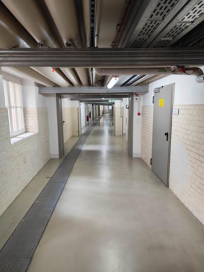 Ein sehr langer, kahler Flur, leciht gekrümmt; unter der Decke Versorgungseinrichtungen (Rohre etc.), rechts eine Tür, links lichtspendende Fenster.