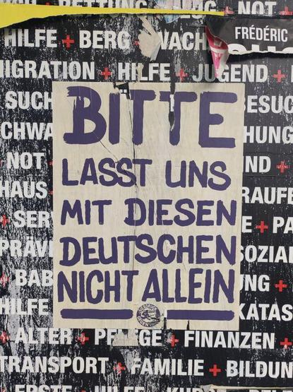 Ein Poster "Bitte lasst uns mit diesen Deutschen nicht allein" mit einem Logo "Siebdruck gegen Nazis" in dem ein Rakel ein Swastika zerstört. Das Poster klebt auf einer Fläche mit schwarzem Hintergrund und weißen Wörtern, wie "Hilfe", "Not", "Finanzen". Das Wort "Naxis" im Logo des Posters wurde mutwillig zerkratzt, ist aber noch zu erahnen.