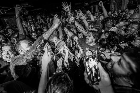 L'immagine è una fotografia in bianco e nero che cattura un momento di intensa energia e partecipazione durante un concerto. La scena è gremita di persone, molte delle quali alzano le braccia in aria, alcune con pugni chiusi, esprimendo entusiasmo e coinvolgimento. Al centro dell'immagine, un artista sta cantando con passione, con un microfono in mano, mentre la folla lo circonda. Alcuni spettatori stanno registrando l'evento con i loro smartphone, evidenziando l'importanza del momento. La folla è composta da persone di diverse età e stili, con alcuni che indossano giacche di jeans e magliette, e altri con capelli lunghi e barbe. L'atmosfera è elettrizzante, con un senso di unità e condivisione tra i presenti. La fotografia è scattata in un ambiente interno, probabilmente un locale di concerto, con luci soffuse che accentuano l'atmosfera vibrante.