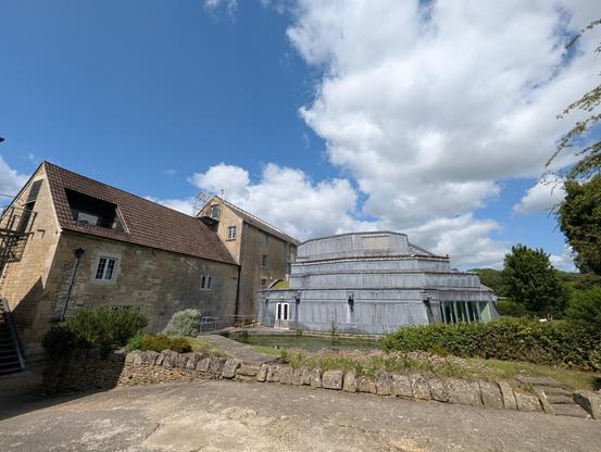 A wide angle shot of the main building at Real World Studios on a sunny day with scattered clouds. The left half of the building is an old mill; the right half is high tech lead-covered concrete and glass. In the foreground is a flower bed with a low brick wall and a paved area.