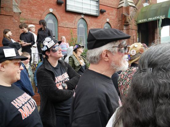 Several molly dancers, most wearing black t shirts, trousers, and hats (one wears a floral print dress) with face paint standing outside a building, watching a performance out of frame to the right. The t shirts say "HANDSOME MOLLY". There are several other audience members around.