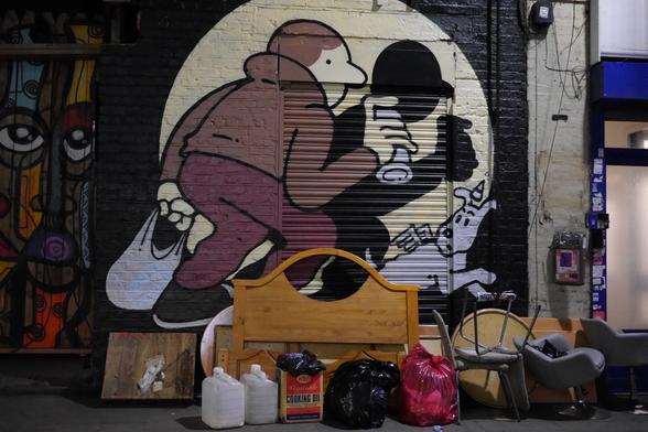 Foto eines großen Graffitis an einer Wand mit Rolladen, davor alleglei Sperrmüll. Dargestellt ist ein Sprayer, der eine Plastiktüte in der einen, eine Spraydose in der anderen Hand hat. Er steht im Spotlight und schaut sich mit hochgeschlagenem Kragen vorsichtig um. Rechts ein weißer Hund mit kleinen Engelsflügeln und einem spitzen Hütchen.