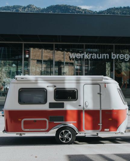 Das Bild zeigt eine kleine, rote und weiße Wohnwagen, der auf einer Straße geparkt ist. Der Wohnwagen hat eine klassische, retroartige Form mit einem schrägen Dach und einer Tür auf der rechten Seite. Die Fenster sind mit Rollläden versehen, und an der Seite befindet sich ein kleiner Schrank. Der Wohnwagen steht vor einem modernen Gebäude mit großen Glasfenstern, auf denen "werkraum bregenz" steht. Im Hintergrund sind bewaldete Hügel zu sehen, was auf eine ländliche oder bergige Umgebung hinweist. Die Sonne scheint, und es ist ein klarer Tag.