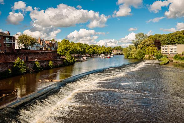 A wide river with a large weir, lined with trees and houses. It is a lovely clear day with a blue sky and fluffy white clouds.
