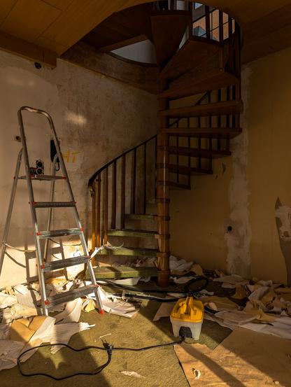 Das Bild zeigt eine Renovierungsszene in einem Innenraum. Es gibt eine Wendeltreppe aus Holz, die nach oben führt. Die Wände zeigen Spuren von alten Tapeten und Putz. Auf dem Boden liegen viele Stücke von abgerissenen Tapeten und anderen Materialien verstreut.

An einer Wand lehnt eine Leiter, und daneben steht ein gelbes Dampf-Gerät zur Tapetenentfernung.
Die Szene wirkt chaotisch und zeigt den Fortschritt der Renovierungsarbeiten.