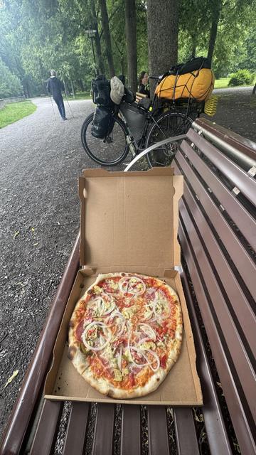 Eine frische Pizza mit Belägen wie Zwiebeln, Paprika und Oliven wird auf eine Bank in einem Park gestellt. Im Hintergrund ist ein Fahrrad geparkt, während zwei Personen auf einem von viel Grün umgebenen Weg in der Nähe gehen.