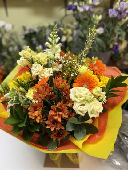 A hand tied bouquet of all mixed yellow and orange flowers including gerbera, carnations, daisy chrysanthemum, lisianthus, altromeria and mixed foliages, wrapped in bright sunshine yellow wrapping with an orange tissue inlay.