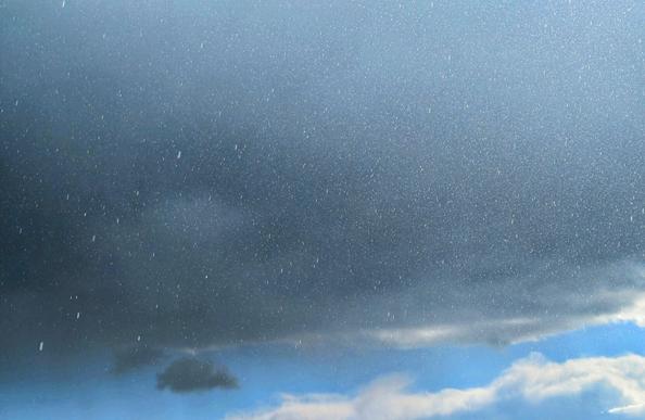 A close-up shot shows a grey, overcast sky with rain falling. The raindrops are visible as small, bright streaks against the dark clouds. Towards the bottom of the frame, lighter, white clouds are visible against a patch of blue sky.