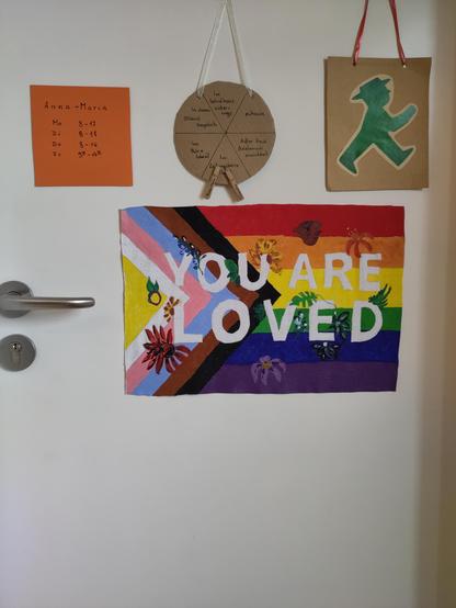 Foto meiner Bürotür: Man sieht meine Sprechzeiten aushängen, eine Klammerkarte mit Wäscheklammern, die anzeigen, wo ich und meine Kollegin gerade im Schulhaus sind. Ein grünes Ampelmännchen zeigt, dass die Tür zum Büro gerade offen steht. 
Unter diesen Dingen hängt eine selbst gemalte Progress-Pride-Flagge mit der Aufschrift "You are loved" und vielen Blumen.