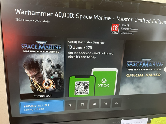 A close-up of a computer monitor displaying the Xbox Game Pass page for "Warhammer 40,000: Space Marine - Master Crafted Edition." The game's title is at the top, along with "SEGA Europe • 2025 • 44GB" and a PEGI 18 rating for "Extreme Violence" and "Users Interact." On the left, the game's cover art shows a grizzled Space Marine with a glowing gauntlet and a spectral skull. In the center, a section reads "Coming soon to Xbox Game Pass 10 June 2025" and "Get the Xbox app - we'll notify you when it's time to play." Below this text is a green image of a mobile phone screen displaying a QR code next to a green Xbox logo. On the right, another game tile shows "Warhammer 40,000: Space Marine Master Crafted Edition OFFICIAL TRAILER." A blue "PRE-INSTALL ALL Coming in 8 days" button is visible at the bottom left.