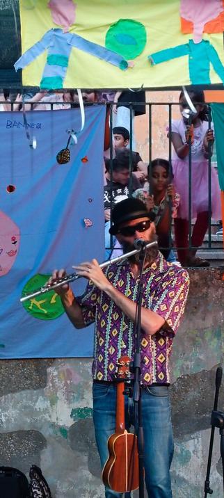 Un musicista suonando il flauto è al centro dell'immagine, indossando una camicia colorata con un motivo geometrico e un cappello nero. Sullo sfondo, un gruppo di persone, tra cui bambini, osserva il concerto da dietro una ringhiera. Sullo sfondo, un cartello blu con disegni colorati e la scritta "BANDERA" è visibile. Un altro cartello con figure umane colorate è appeso sopra. Il musicista ha un microfono davanti a sé e un chitarra è appoggiata al suo fianco. L'atmosfera sembra festosa e informale, con un'atmosfera di comunità.