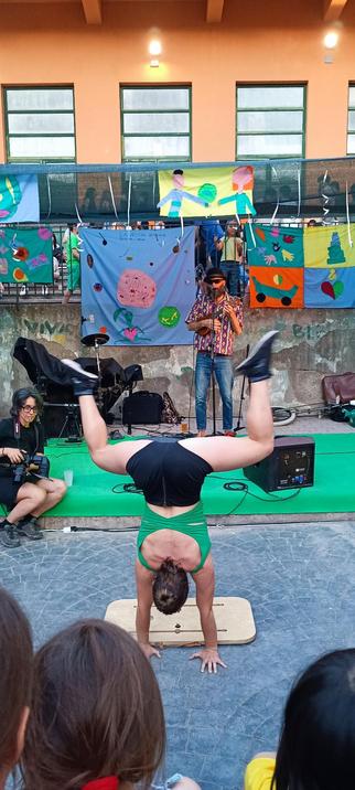 L'immagine mostra una performance artistica all'aperto. Al centro, una persona esegue un handstand su una pala da yoga, indossando un top verde e pantaloncini neri. Sullo sfondo, un musicista suona un ukulele, mentre un fotografo cattura l'evento. Sullo sfondo, un palco è decorato con bandiere colorate e disegni creativi, suggerendo un contesto festivo o culturale. La scena è illuminata da luci artificiali, e il pavimento è di pietra.