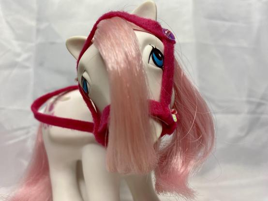 A toy horse with a pink mane and eyes, wearing a bright pink harness. The background is a plain white surface, highlighting the horse's features.