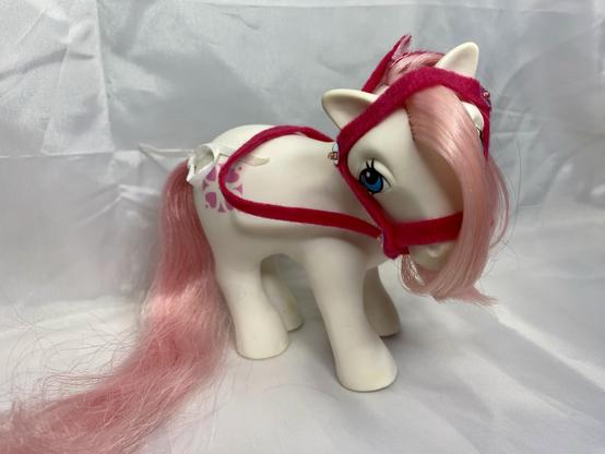 A toy pony with a white body and pink mane. It is wearing a red harness and has a decorative symbol on its side. The background is a soft, white fabric.