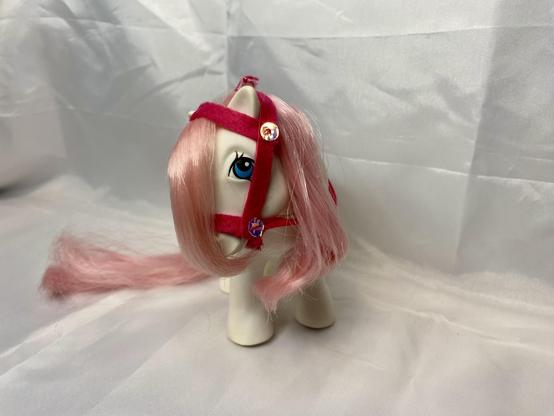 A toy pony with pink hair and a red harness, featuring large blue eyes. The background is a simple white fabric.