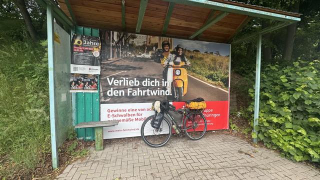 Ein Fahrrad mit Taschen lehnt sich an einen Bushaltestellenschutz. Es gibt eine Mini-Bank für maximal eine Person.