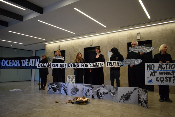 Candles lit in front of pictures showing dead sea animals as a result of the algal blooms. People standing behind holding signs including “ocean kin are dying for climate truth”